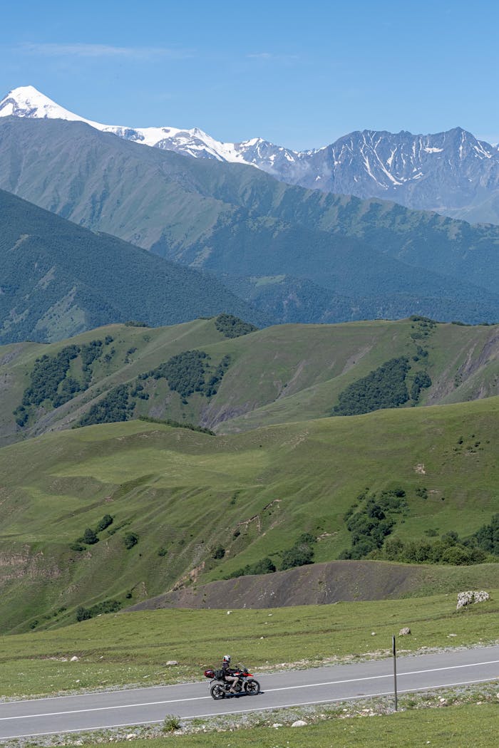 A motorcycle rides through a lush green road with snow-capped mountains in the background.